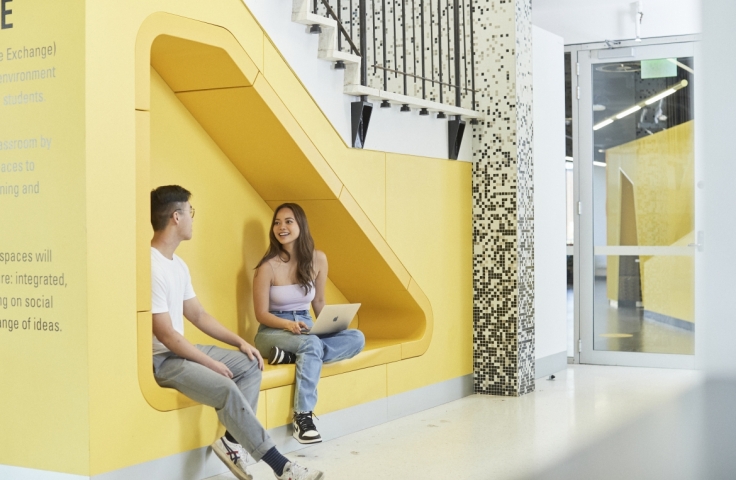 Two student sitting with laptops against a yellow wall